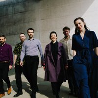 Sechs Personen, lässig gekleidet, gehen im sonnigen Wetter als Gruppe vor der Betonfassade der Bonner Bundeskunsthalle.