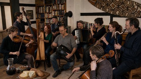 Eine Gruppe von Musikern spielt eine Vielzahl von Instrumenten in einem gemütlichen Raum. Brot und Wein auf dem Tisch.