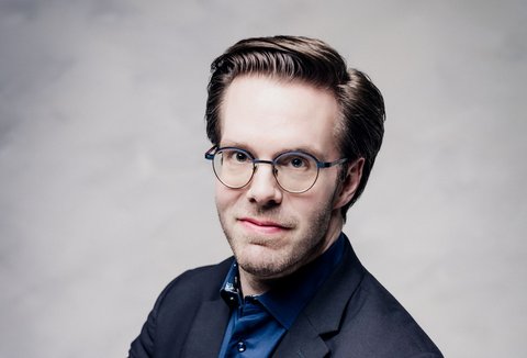 Ein Mann sitzt vor einem hellen Hintergrund. Er trägt einen Anzug und hat die Arme verschränkt. Er blickt in die Kamera.