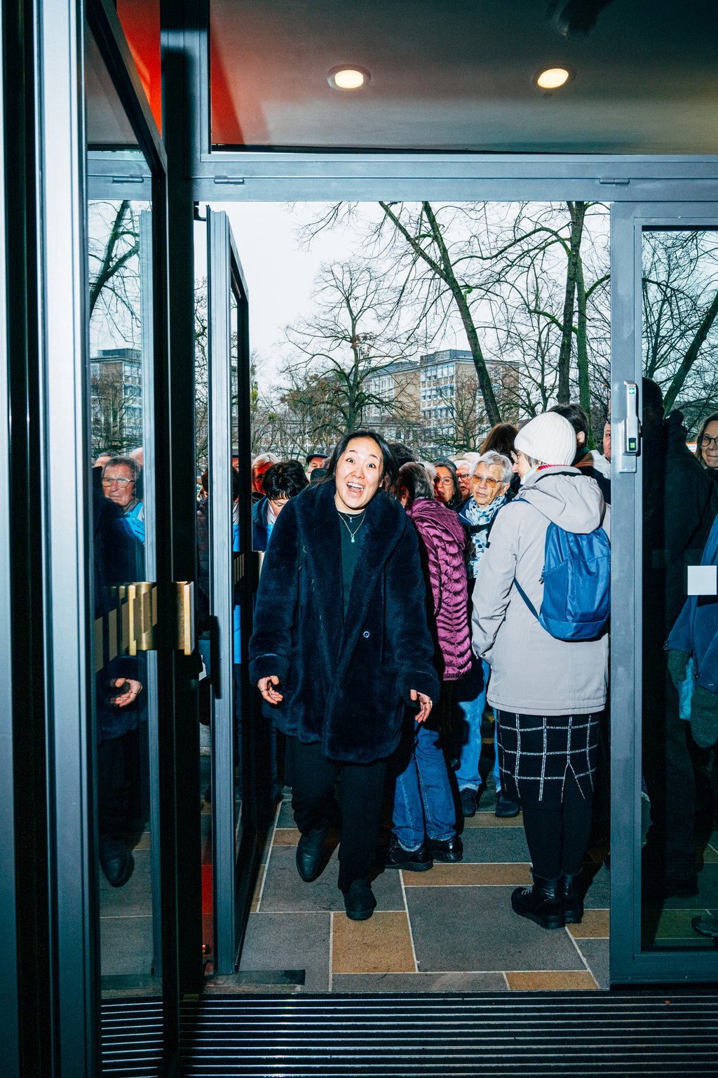 Eine Person in einem schwarzen Mantel betritt lächelnd ein Gebäude. Eine Gruppe von Menschen folgt ihr durch die Glastür.
