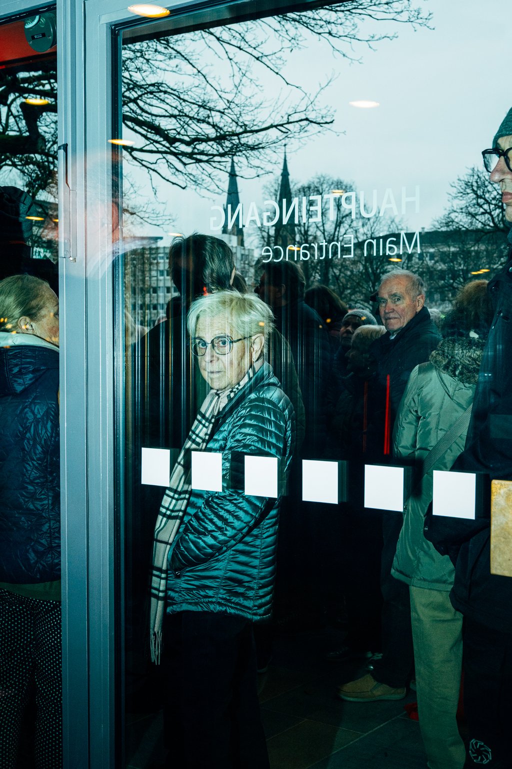 Menschen warten in einer Schlange an einem Eingang mit Glastür. Ältere Person mit Brille schaut durch die Tür.