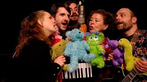 Fünf Personen singen in ein Mikrofon, umgeben von bunten Plüschtieren. Die fröhliche Gruppe wirkt in einem musikalischen Auftritt.  © Claus Overkamp
