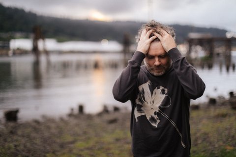 Ein Mann steht vot einem See auf einer Wiese, er trägt einen dunklen Pulli mit einer großen aufgedruckten Blüte in weiß. Er hat seine Hände in seine grauen Locken vergraben und schaut nach unten. Die Stimmung ist düster.  © Jason Quigley