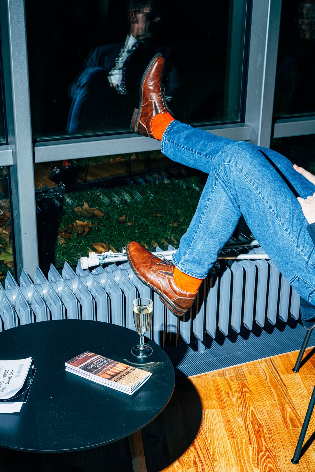 Person mit blauen Jeans und orangenen Socken, Füße auf der Fensterbank, Glas Sekt und Bücher auf dem Tisch. Man sieht nur die Beine.