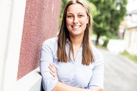 Eine Person in einem hellblauen Hemd lächelt und steht vor einer rot gestrichenen Wand. Im Hintergrund ist ein Weg mit Bäumen zu sehen.  © unbezeichnet