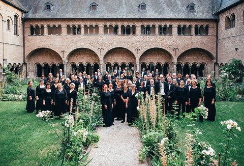 Ein Chor posiert in einem historischen Innenhof mit Bögen, umgeben von üppigen Gärten und Blumen in voller Blüte.