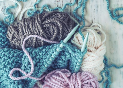 Verschiedene Wollgarne in Blau, Lila und Weiß neben Stricknadeln auf einem Holztisch. Unvollendetes Strickprojekt mit texturiertem Muster.
