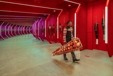 Person in einem futuristischen, roten Gang trägt ein Balafon, ein westafrikanisches hölzernes Xylophon mit runden Resonanzkörpern. Die Beleuchtung ist in violetten und roten Tönen gehalten.