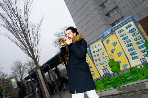 Ein Mann spielt Trompete vor einer farbenfrohen Street Art auf einemStromkasten, die ein Wohngebäude zeigt. Im Hintergrund ist ein graues Hochhaus und eine Bushaltestelle. Der Himmel ist bewölkt.