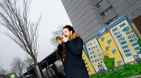 Ein Mann spielt Trompete vor einer farbenfrohen Street Art auf einemStromkasten, die ein Wohngebäude zeigt. Im Hintergrund ist ein graues Hochhaus und eine Bushaltestelle. Der Himmel ist bewölkt.