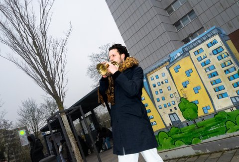 Ein Mann spielt Trompete vor einer farbenfrohen Street Art auf einemStromkasten, die ein Wohngebäude zeigt. Im Hintergrund ist ein graues Hochhaus und eine Bushaltestelle. Der Himmel ist bewölkt.