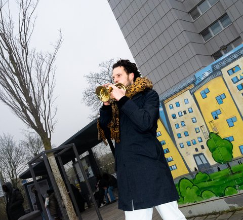 Ein Mann spielt Trompete vor einer farbenfrohen Street Art auf einemStromkasten, die ein Wohngebäude zeigt. Im Hintergrund ist ein graues Hochhaus und eine Bushaltestelle. Der Himmel ist bewölkt.