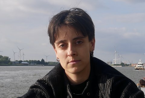 Eine Frau sitzt vor einem Fluss. Sie trägt eine dunkle Jacke. Sie blickt in die Kamera.