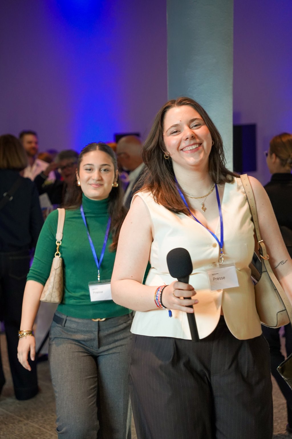 Zwei Frauen bei einer Veranstaltung. Eine hält ein Mikrofon, beide lächeln. Im Hintergrund sind andere Teilnehmer zu sehen.