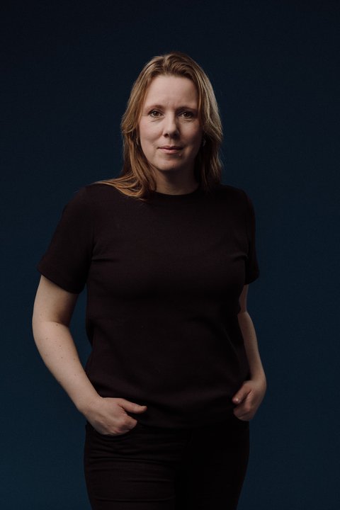 Ein stehendes Porträt einer Frau mit halblangen blonden Haaren. Sie hat ihre Hände in die Hosentaschen gesteckt und lächelt entspannt. Sie trägt schwarze Kleidung. Der Hintergrund ist schwarz.  © Marius Fiskum