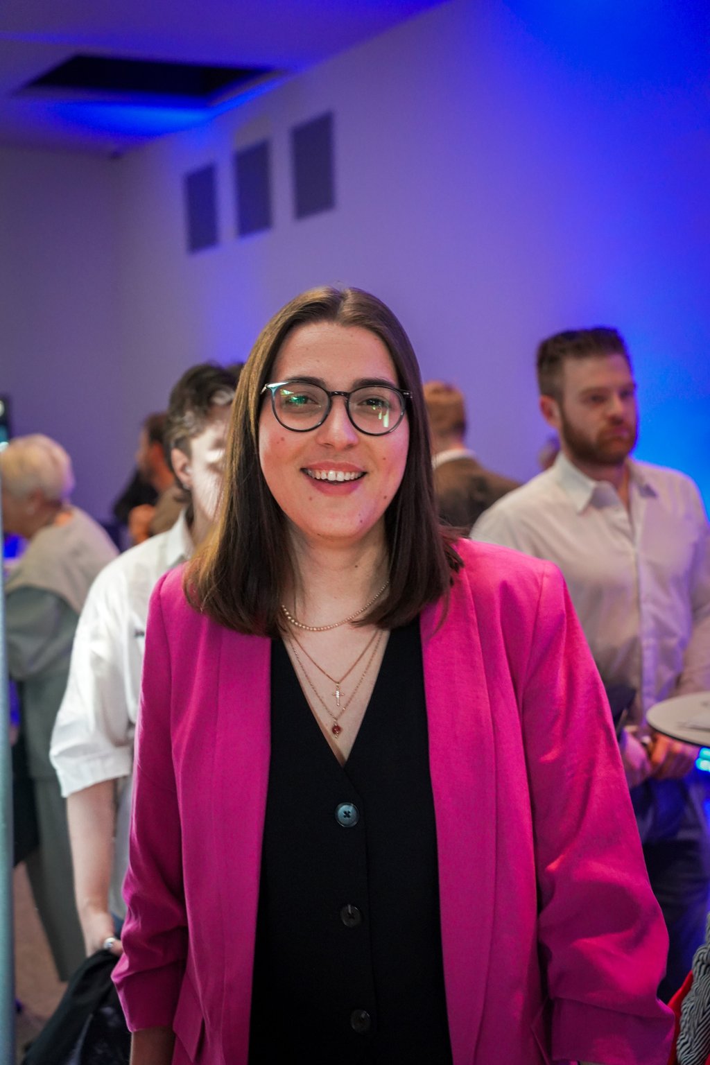 Junge Frau in einem pinken Blazer lächelt bei einer Veranstaltung. Im Hintergrund sind weitere Gäste und blaues Licht.