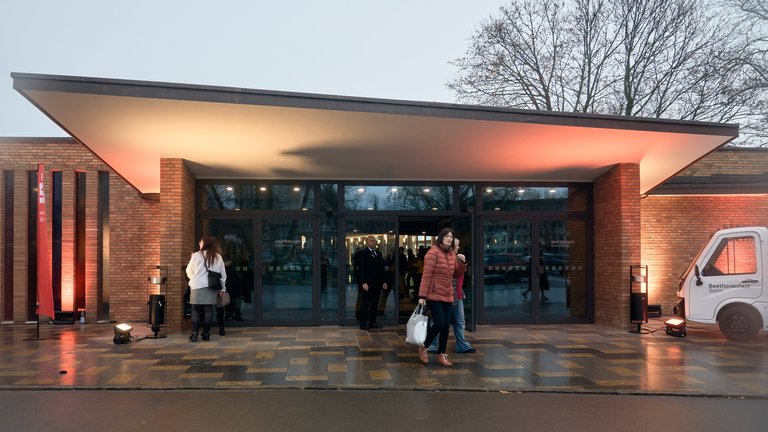 Personen betreten ein modernes Gebäude mit Glasfassade, abends beleuchtet, um ein Event zu besuchen. Bäume sind im Hintergrund zu sehen.