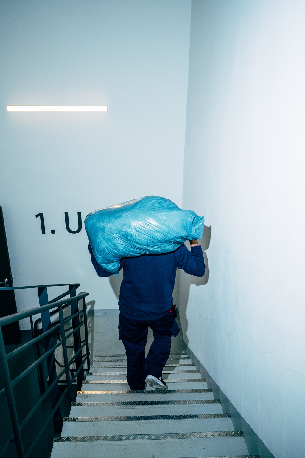Eine Person trägt einen großen blauen Sack die Treppe hinauf, in einem modernen Gebäudeflur. Es ist eine helle Umgebung.