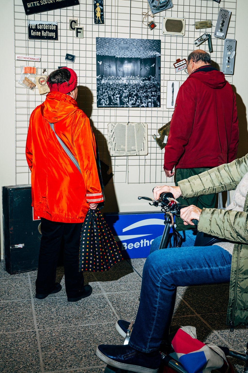 Personen betrachten die Wandcollage mit Bildern und Objekten im Ausstellungsraum der Beethovenhalle. Eine Person im Rollstuhl ist im Vordergrund zu sehen.