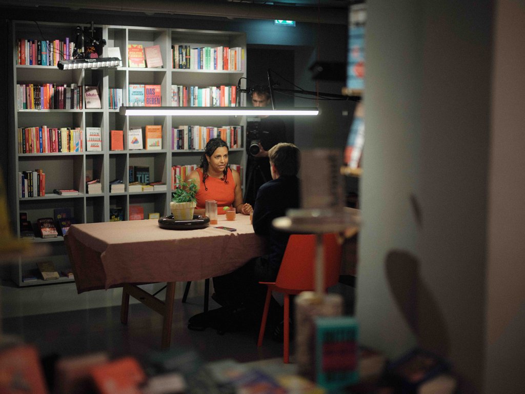 Eine Frau in einem roten Oberteil und ein Mann in einem dunklen Hemd sitzen in einer Buchhandlung. Sie unterhalten sich und werden gefilmt.