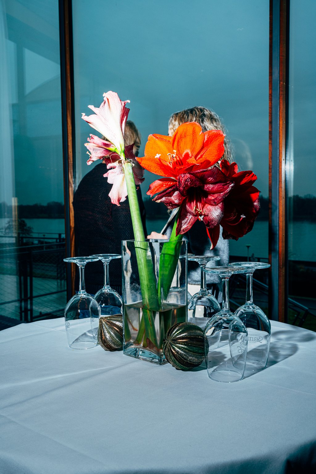 Ein Blumenarrangement mit Amaryllis-Blüten steht auf einem gedeckten Tisch, umgeben von leeren Weingläsern, vor einem Fenster. Die Blumen verdecken 2 Frauen dahinter.