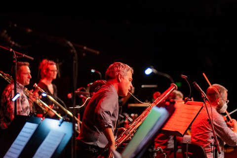 Eine Gruppe von Musikern spielt auf der Bühne Blasinstrumente. Sie sind in rot beleuchtet, während sie konzentriert lesen und spielen.