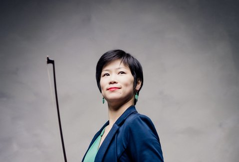 Eine Frau mit einer Violine steht vor einem hellen Hintergrund. Sie trägt einen Anzug und lächelt.