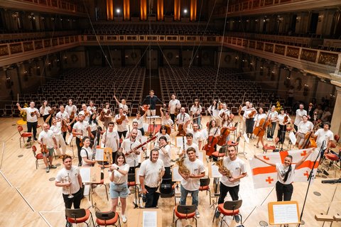 Eine große Gruppe von Orchestermusikern steht auf einer Bühne. Sie tragen weiße Hemden und halten verschiedene Instrumente.  © privat