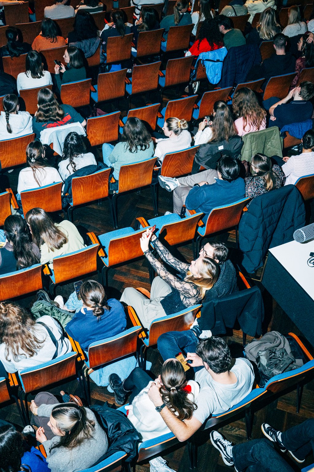 Menschenmenge in einem Auditorium mit jungen Zuhörern. Ein Paar ein Selfie macht. Holzstühle mit blauem Bezug füllen den Raum.