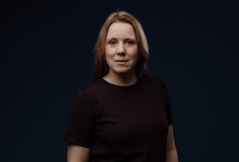 Eine Frau im Porträt, sie hat halblange dunkelblonde Haare und trägt ein dunkles Oberteil. Sie schaut neutral lächelnd nach vorn. Der Hintergrun ist schwarz.