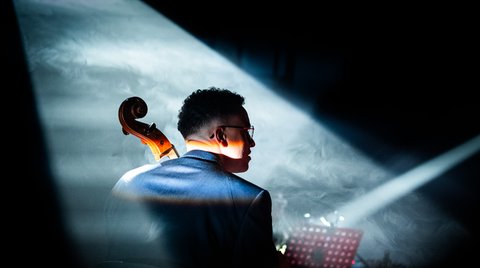 Ein Mann spielt das Violoncello in einem Konzert. Sein Rücken ist zur Kamera, und sein Kopf ist nach rechts gedreht, sodass sein Gesicht im Profil ist.