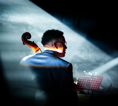 Ein Mann spielt das Violoncello in einem Konzert. Sein Rücken ist zur Kamera, und sein Kopf ist nach rechts gedreht, sodass sein Gesicht im Profil ist.
