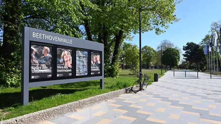 Plakatwand vor der Beethovenhalle mit Werbeplakaten. Ein sonniger Pfad, gesäumt von Bäumen, führt in die Ferne.