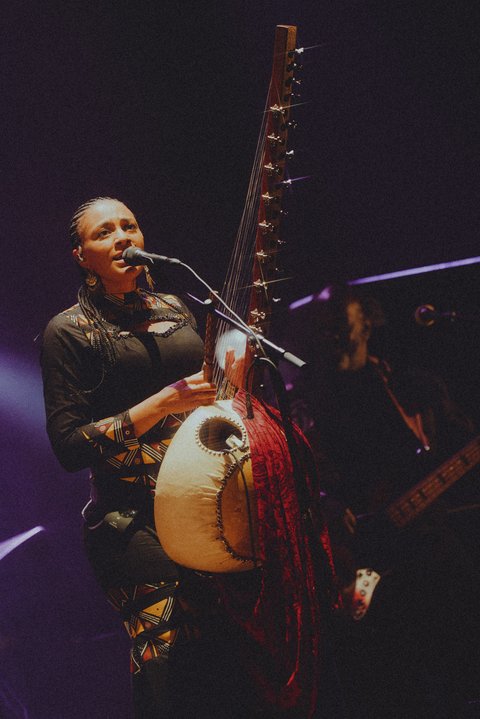 Eine Musikerin spielt eine Kora und singt in ein Mikrofon auf einer Bühne. Die Beleuchtung ist dunkel und stimmungsvoll.  © Daria Miasoedova