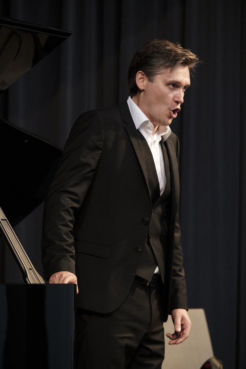 Ein Sänger steht an einem Flügel und singt. Er trägt einen schwarzen Anzug und ein weißes Hemd. Er schaut intensiv nach vorne. Der Hintergrund ist schwarz.  © Niclas Weber