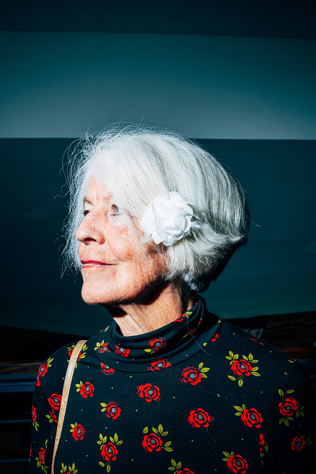 Eine ältere Frau mit weißen Haaren und einer Blume im Haar. Sie trägt einen schwarzen Pullover mit roten Rosen.