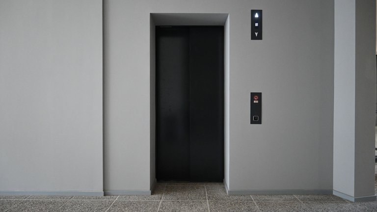 Ein moderner Aufzug in einem Gebäude. Die offenen Türen zeigen einen minimalistischen Stil mit grauen Wänden und steinartigem Boden.