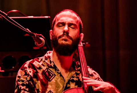 Ein Musiker spielt ein Cello auf einer Bühne mit melodischer Leidenschaft. Der Hintergrund ist stimmungsvoll rot beleuchtet.