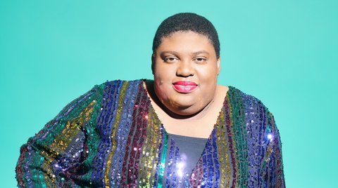 Eine Person in einem glänzenden Regenbogen-Glitzerkleid sitzt auf einem türkisfarbenen Hintergrund und lächelt freundlich in die Kamera.