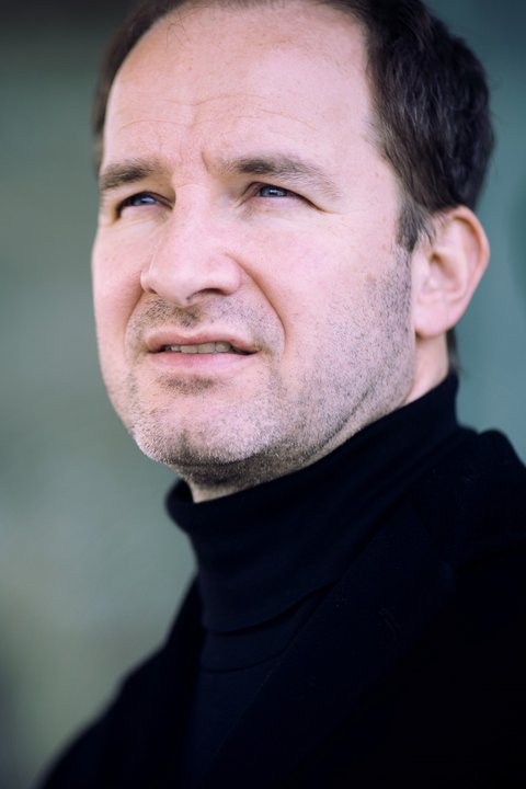 Ein Mann im Porträt, er schaut ernst an der Kamera vorbei. Er trägt einen schwarzen Rollkragenpullover. Der Hintergrund ist türkis-grau.  © anna.s