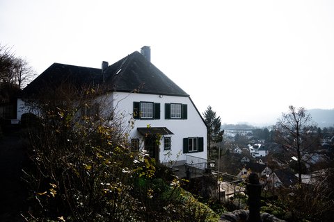 Ein weißes Haus mit dunklen Fensterläden steht auf einem Hügel mit Blick auf eine Landschaft und Stadt.