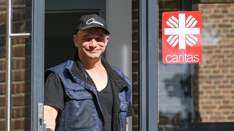 Ein Mann mit schwarzer Kappe und Weste steht lächelnd an einer Tür. Ein Caritas-Schild ist sichtbar.