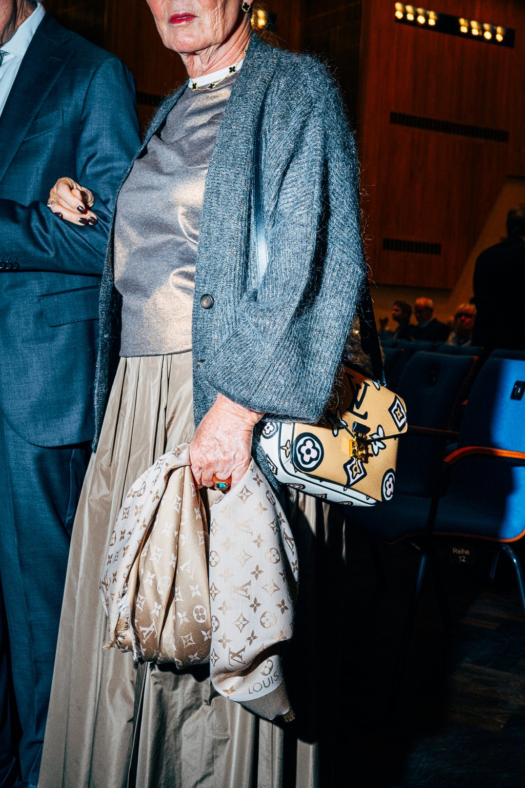 Eine Frau in eleganter Kleidung mit grauer Jacke und Rock hält eine modische, verzierte Tasche bei einer Veranstaltung.