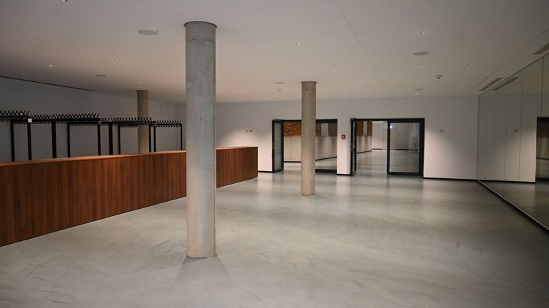 Leerer Raum mit grauem Boden, hölzerner Theke und Säulen. Große Fensterfront rechts, moderne und minimalistische Gestaltung.