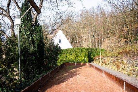 Ein Sandboden, eingezäunt von Metallbarrieren, führt zu einem weißen Gebäude im Hintergrund. Bäume ohne Blätter umgeben die Szene.