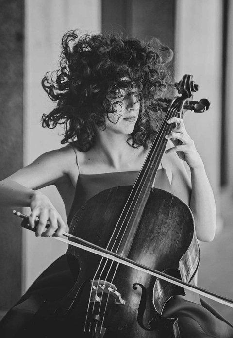 Schwarz-weiß Foto einer Frau, die ein Cello mit leidenschaftlichem Ausdruck spielt. Ihr lockiges Haar bewegt sich dynamisch.