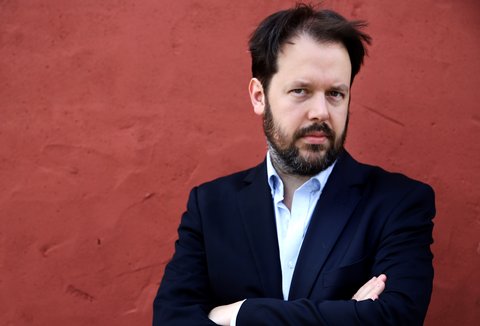 Ein Mann steht vor einem farbigen Hintegrund. Er trägt einen Anzug. Seine Arme sind verschränkt. Er blickt in die Kamera.