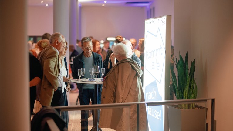 Menschen stehen in einer elegant beleuchteten Galerie oder Veranstaltungshalle, unterhalten sich und genießen Getränke. Pflanzen und Schilder sind sichtbar.