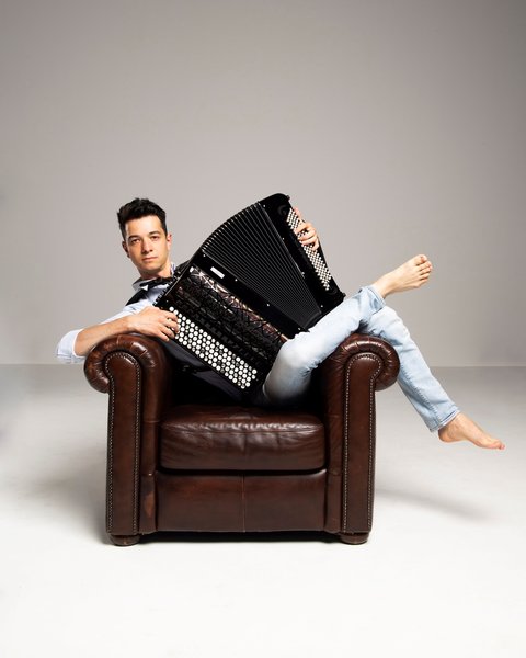 Ein Mann posiert mit seinem Akkordeon vor einem hellen Hintergrund. Er streckt sich barfuß in einem Sessel und blickt in die Kamera.  © Pigini Accordions