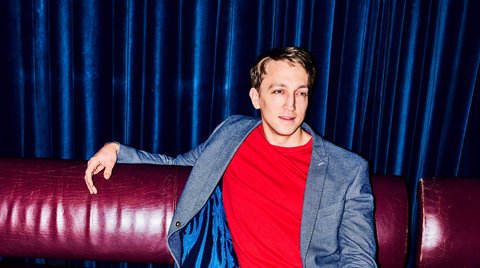 Ein Mann in blauem Blazer und rotem T-Shirt sitzt entspannt auf einem roten Sofa mit einem Drink auf dem Tisch.
